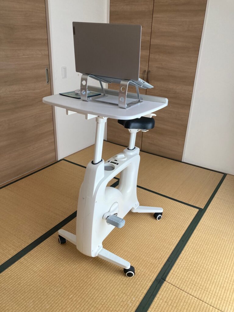 デスク付きエアロバイクにノートパソコンを置き、ペダルとAI管理人えりりがブログ記事を作成している実際の作業環境。